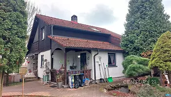 Rodinný dům se šikmou střechou a cihlovými taškami, obložený dřevem. Prostorná zahrada s chodníkem a okrasnými stromy.