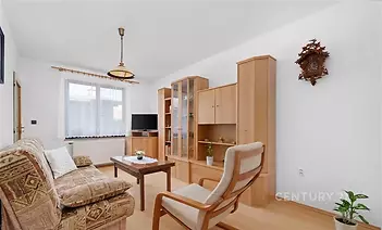 Světlý obývací pokoj s parketovou podlahou, velkým oknem a dřevěným nábytkem, který dotváří moderní a útulný vzhled.