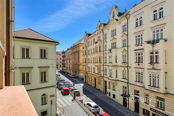 Pohled z balkonu na historické budovy s dekorativními fasádami, klidná dlážděná ulice, ideální městská lokalita pro bydlení.