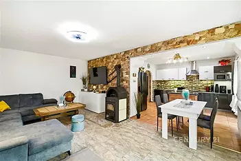 Stylový obývací pokoj s krbovými kamny a parketovou podlahou, propojený s moderní kuchyní. Velká okna zajišťují dostatek světla.