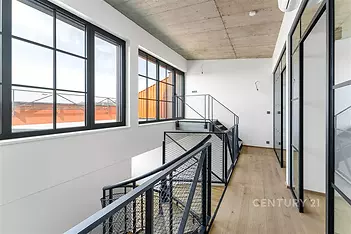 Světlá chodba s velkými okny, moderním průmyslovým stylem a dřevěnou podlahou. Skleněné stěny s kovovými rámy dodávají industriální vzhled.