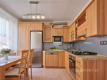 Moderní kuchyň s dřevěnou linkou, dlažbou na podlaze a vestavěnými spotřebiči, prostorná a světlá díky velkým oknům.