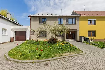 Rodinný dům s tradiční fasádou, upravenou zahradou a garáží. Přístupová cesta je z dlažby. Dům má moderní okna a udržovaný exteriér.