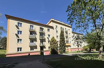 Čtyřpatrový bytový dům s balkony, okna s bílými rámy, obklopen zelení, v klidné rezidenční čtvrti, dobrý stav, cihlová fasáda.