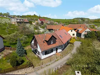 Rodinný dům s červenou střechou a dřevěným balkonem, obklopený zahradou, v klidné vesnické lokalitě s výhledem na okolní kopce.