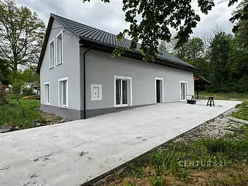 Moderní rodinný dům s šedou fasádou a tmavou střechou, obklopený zelení. Venkovní prostor zahrnuje betonovou terasu, ideální pro relaxaci.