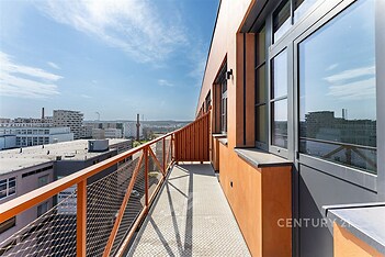 Prostorný balkon s kovovým zábradlím, velká plastová okna, moderní design, s výhledem na městskou zástavbu a jasnou oblohu.