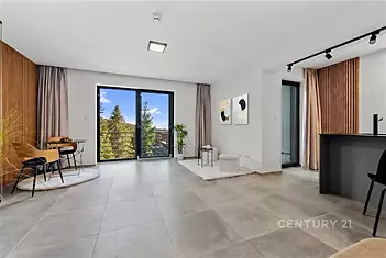 Světlý obývací pokoj s velkými okny, dlažbou a moderním designem, propojený s kuchyňským koutem, nabízí výhled na přírodu.