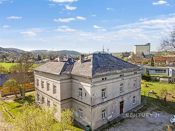 Historická budova s šedou fasádou, umístěná v klidném prostředí, obklopená zelení, ideální pro rekonstrukci nebo komerční využití