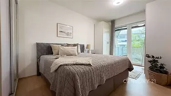 Světlá ložnice s moderním stylem, parketová podlaha, velká okna vedoucí na balkon, vestavěné skříně poskytující dostatek úložného prostoru.