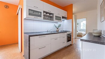 Moderní kuchyň s bílými skříňkami, světlými dlaždicemi a korkovou podlahou, propojená s otevřeným obývacím prostorem a ložnicí.