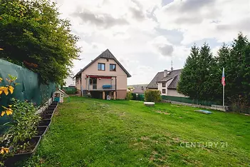 Prostorná zahrada rodinného domu nabízí udržovaný trávník, zelené oplocení a výhled na dům s balkonem; působí svěže a klidně