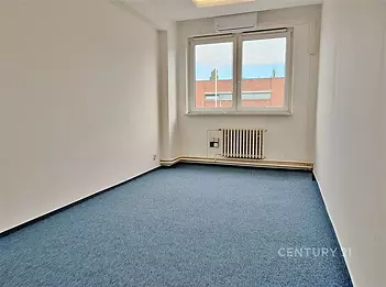 Prostorná světlá místnost s modrým kobercovým povrchem, velkým oknem a bílými stěnami, nabízí moderní kancelářské prostory.