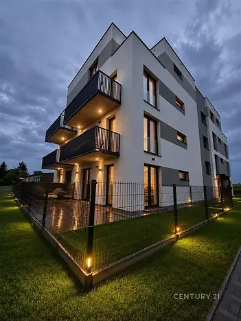Moderní bytová budova s balkony, osvětlená večer, s oplocenou předzahrádkou a pečlivě udržovaným trávníkem