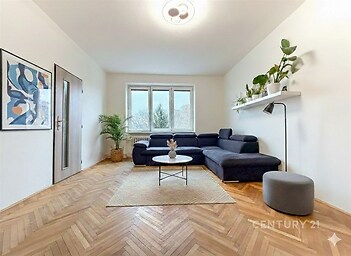 Světlý obývací pokoj s parketovou podlahou, velkými okny a moderním nábytkem, doplněný o dekorativní rostliny a minimalistický styl.