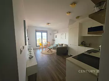 Malý obývací pokoj s dřevěnou podlahou, moderní kuchyní a velkými okny, které poskytují přirozené světlo.