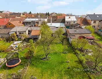 Zahrada s udržovaným trávníkem, vzrostlými stromy a trampolínou, obklopená zděnými domy s různorodou architekturou ve vesnickém prostředí.