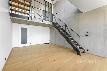 Světlý obývací pokoj s parketovou podlahou, moderní kovové schodiště, betonové stěny a velká okna poskytující přirozené světlo.