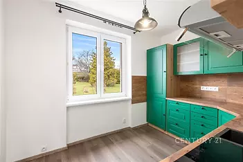 Kuchyň s moderním designem, dřevěnou podlahou a zelenými skříňkami, velké okno zajišťuje světlost a výhled na zahradu.