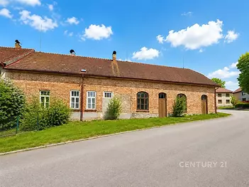 Historická budova s cihlovou fasádou a šikmou střechou, obklopená zelení na klidné ulici, ideální pro přestavbu nebo renovaci.