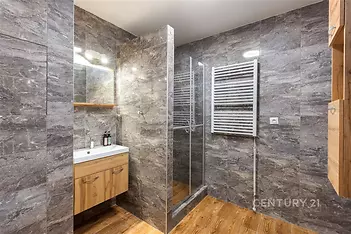 Moderní koupelna s dlažbou, sprchovým koutem a vestavěným umyvadlem. Dřevěná skříňka a světlé osvětlení dodávají elegantní vzhled.