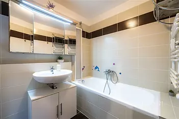 Koupelna s moderními obklady, vanou a velkým zrcadlem, bílé keramické prvky, světlé osvětlení, čistý a udržovaný prostor.