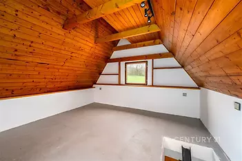 Podkrovní místnost s dřevěným obložením, velkým oknem a betonovou podlahou nabízí světlý a rustikální prostor vhodný pro úpravy.