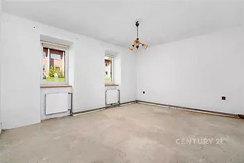 Prázdná světlá místnost, betonová podlaha, bílá výmalba, dvě okna s výhledem na zeleň, příprava na rekonstrukci, ideální pro modernizaci