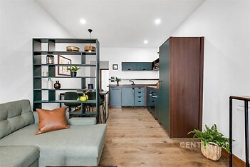 Moderní obývací pokoj s parketovou podlahou, otevřeným konceptem a velkými okny propojený s elegantní kuchyní v tmavém designu.