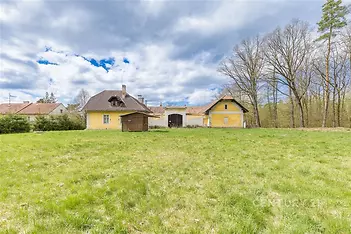 Rozlehlá zahrada s trávníkem obklopujícími venkovský dům, který má šikmou střechu a žlutou fasádu, v klidné lokalitě poblíž lesa.