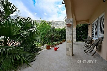 Zahradní terasa s dlažbou, obklopená bujnou zelení, ideální pro relaxaci. Kamenné detaily a přenosné židle vytvářejí příjemnou atmosféru.