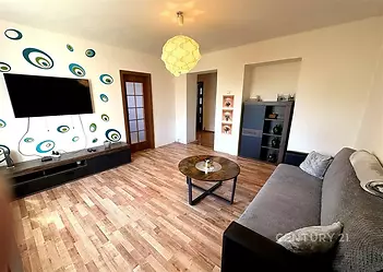 Obývací pokoj s parketovou podlahou, moderní dekorací a světlým nábytkem. Velká okna poskytují dostatek denního světla pro pohodlné bydlení.