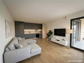 Světlý obývací pokoj s moderní kuchyní, parketová podlaha a velká okna, nabízí otevřený prostor v moderním stylu.