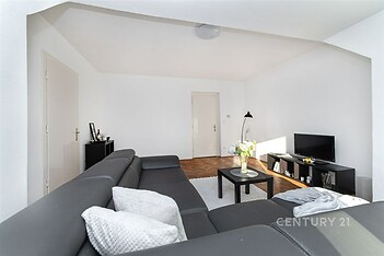 Světlý obývací pokoj s parketovou podlahou, moderní sedačka, velká okna, minimalistický design, černý nábytek, útulný prostor.