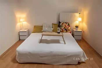 Útulná ložnice, dřevěná podlaha, moderní osvětlení, světlý styl, pohodlná postel, noční stolky, příjemná atmosféra pro relaxaci.