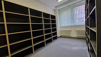 Prostorná kancelář s kobercem, velká okna zajišťují dostatek světla, vestavěné police nabízejí úložné možnosti, moderní interiér.