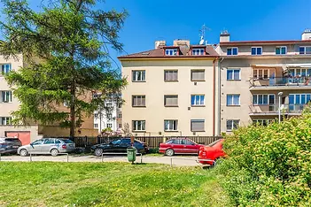 Exteriér bytového domu s udržovanou zahradou, parkováním a červeným autem; ideální lokalita s klidným okolím, vhodná pro rodiny.