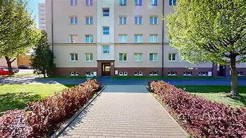 Venkovní fasáda bytového domu se světlou omítkou, upraveným přístupovým chodníkem a okolní zelení, vhodné pro městské bydlení.