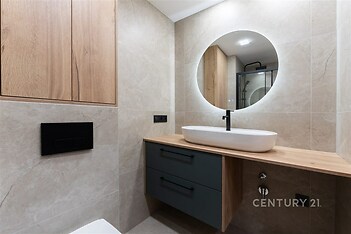 Moderní koupelna s obkladem z dlaždic, kruhovým zrcadlem a dřevěným povrchem. Elegantní umyvadlo podsvícené LED osvětlením.