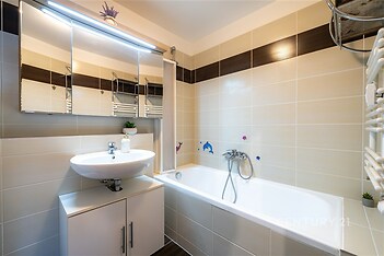 Koupelna s moderními obklady, vanou a velkým zrcadlem, bílé keramické prvky, světlé osvětlení, čistý a udržovaný prostor.
