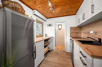Kompaktní kuchyň s moderními spotřebiči, dřevěným obkladem a světlými skříňkami. Dřevěná podlaha dodává příjemnou atmosféru.
