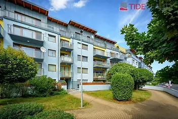Moderní bytový dům s velkými okny a balkony, obklopený upravenou zahradou a parkovou cestou, skvěle situovaný v klidné lokalitě.