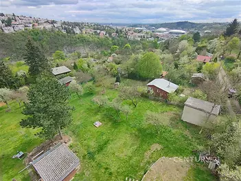Přírodní pozemek s výhledem na krajinu, obklopen stromy a keři, v klidné lokalitě s rekreačními chatami a zelení, ideální pro milovníky přírody.