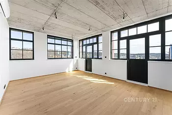 Světlý obývací pokoj s parketovou podlahou, velkými okny a moderním industriálním stylem, ideální pro široké využití.