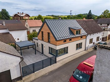 Moderní rodinný dům s dřevěným obložením, velká okna, střecha s plechovou krytinou, terasa s dlážděným povrchem, klidná ulice v rezidenční oblasti.