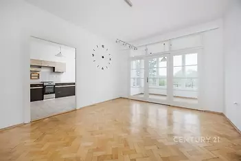 Prostorný obývací pokoj s parketovou podlahou a velkými okny, světlý interiér a moderní kuchyň s dlažbou v pozadí.