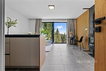 Moderní obývací pokoj s velkými okny, keramická dlažba a dřevěný obklad stěn, výhled na přírodu, světlý interiér s minimalistickým designem.