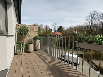 Prostorný balkon s dřevěnou podlahou, moderním zábradlím a rostlinami v květináčích, nabízí výhled na klidné okolí.