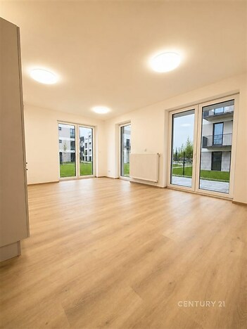 Světlý obývací pokoj s parketovou podlahou, velká okna poskytující výhled do zahrady, moderní design a světlé stěny.