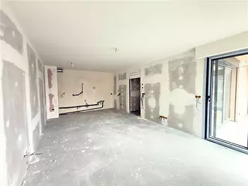 Prázdná místnost s betonovou podlahou, velkými okny a přípravou na moderní kuchyň. Prostor ideální pro rekonstrukci dle vlastních představ.